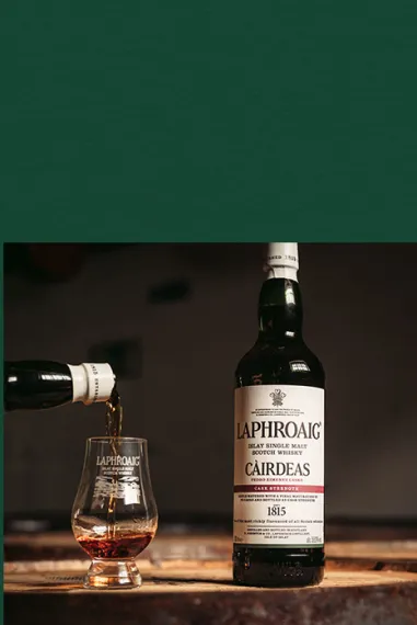 A picture of a Cairdeas Laphroaig bottle with a dram being filled beside it, on dark green background