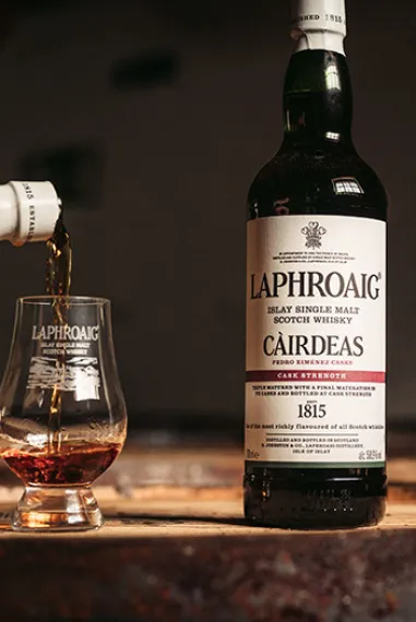 A bottle of Cairdeas Laphroaig standing on a barrel with a dram beside it that is being filled, and the Friends of Laphroaig Emblem put beside the bottle