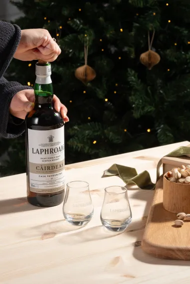 A person opening a Laphroaig Cairdeas 2024 bottle on a festive set table next to two nosing glasses with a Christmas tree in the background
