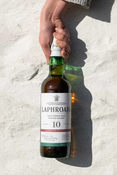 Hand holding a bottle of Laphroaig 10 Year Old Sherry Oak Cask Finish Islay single malt Scotch whisky against a white wall.