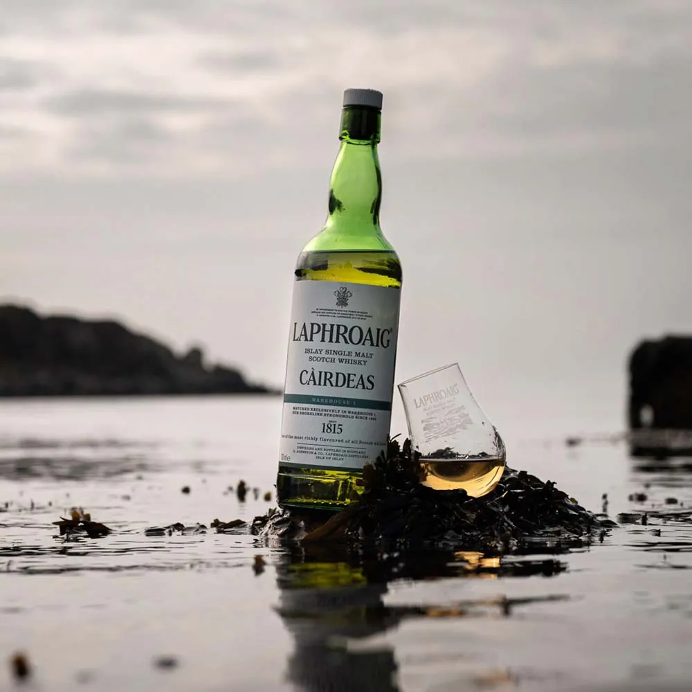 Bottle of Laphroaig Cairdeas Warehouse 1 with dram of whisky on seaweed on an overcast beach