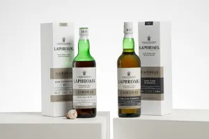 Two bottles of Laphroaig Càirdeas Islay single malt Scotch whisky displayed with their matching presentation boxes on white plinths against a minimalist white background. The bottle on the left is Cask Favourites 10, while the bottle on the right is Lore Cask Strength. A small cork rests beside the left bottle.