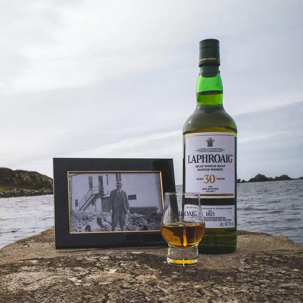 Whisky heritage on the Islay shore: A Laphroaig bottle standing on a rock with a filled dram and a photo of the Laphroaig distillery founder beside it