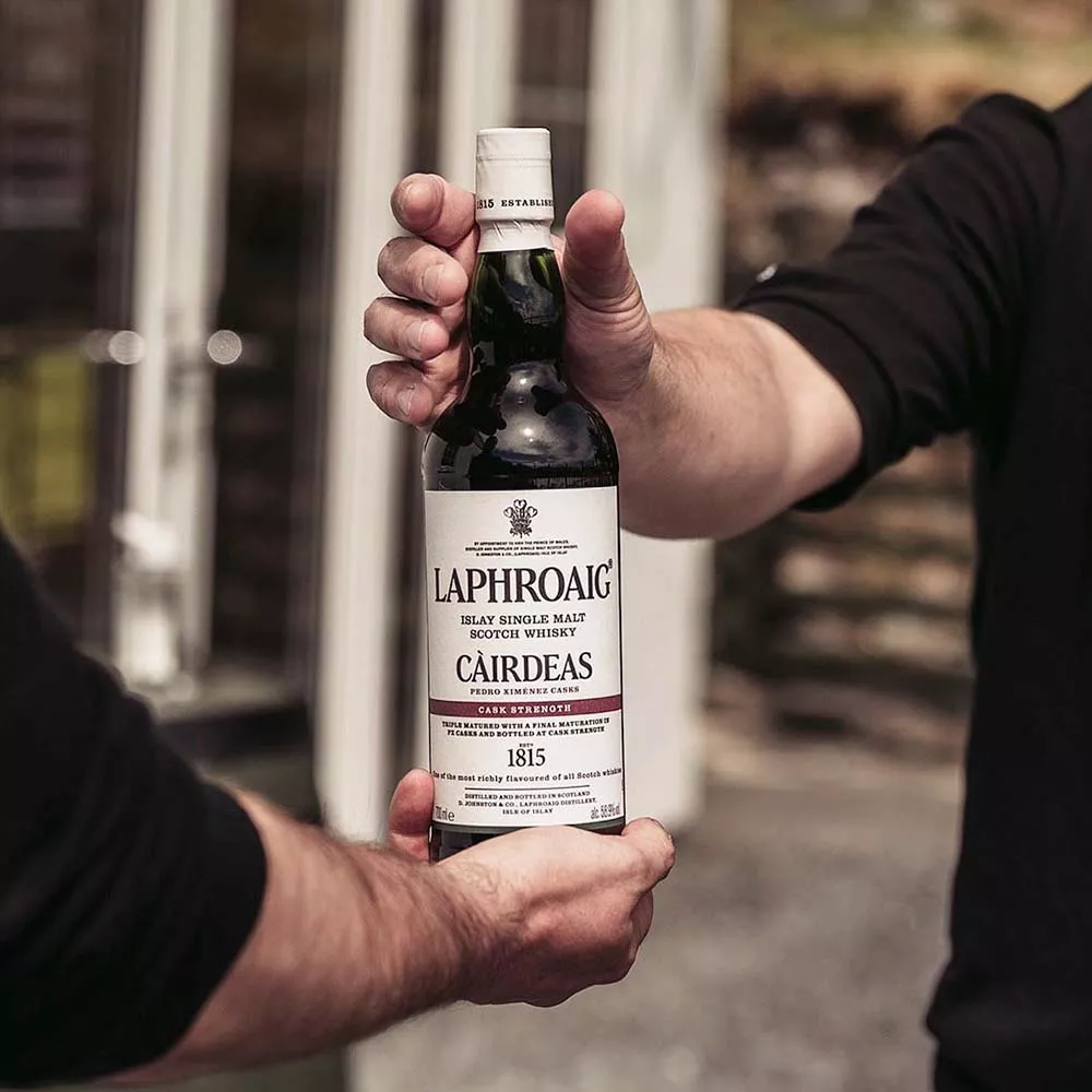 One hand holding Laphroaig Cairdeas 2021 and one hand reaching out to grab bottle