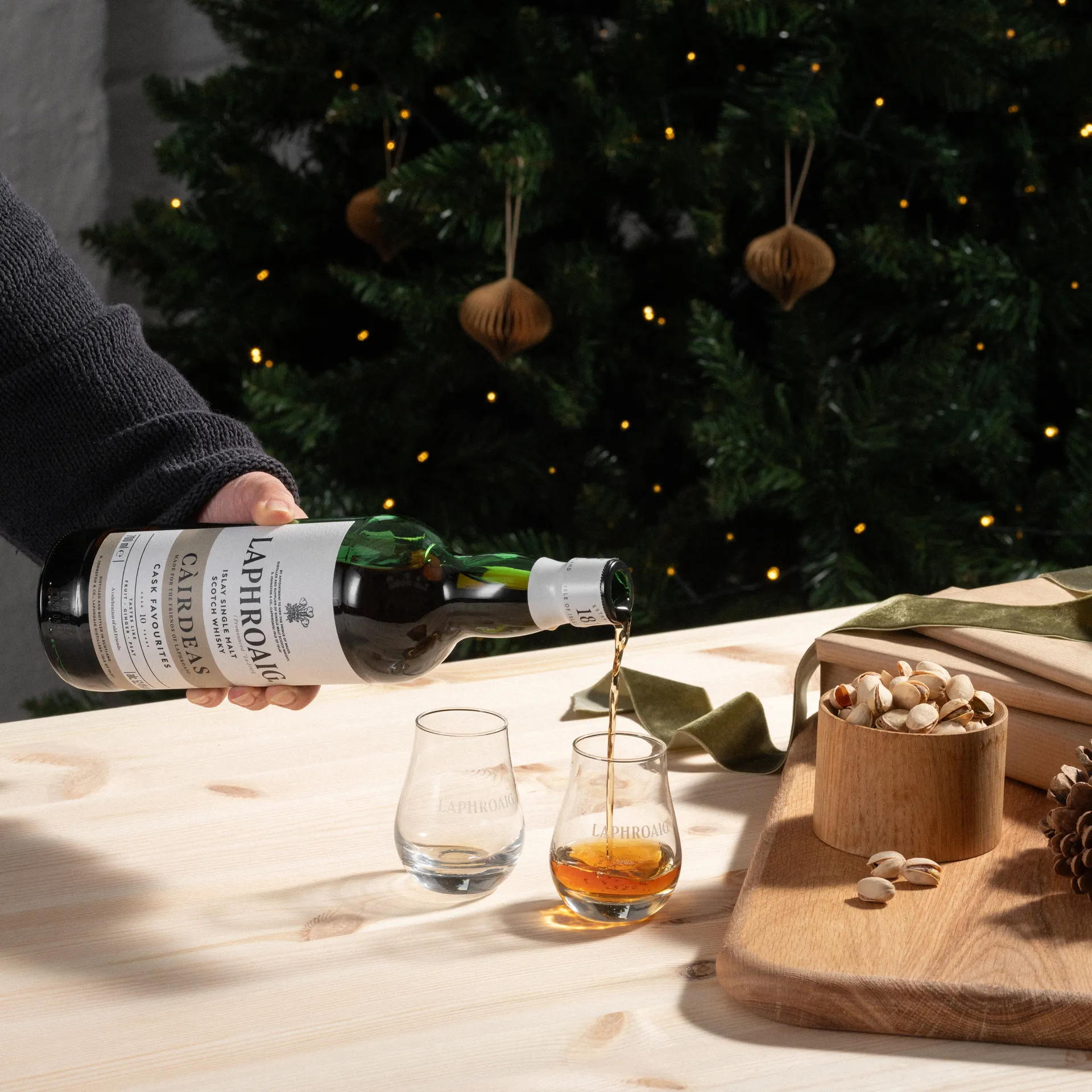 A person pouring a Cairdeas dram into a nosing glass on a festive set table with a Christmas tree in the background