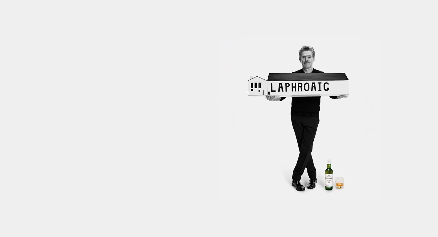 Willem Dafoe holding a miniature Laphroaig distillery with a Laphroaig 10 on the floor next to a poured dram