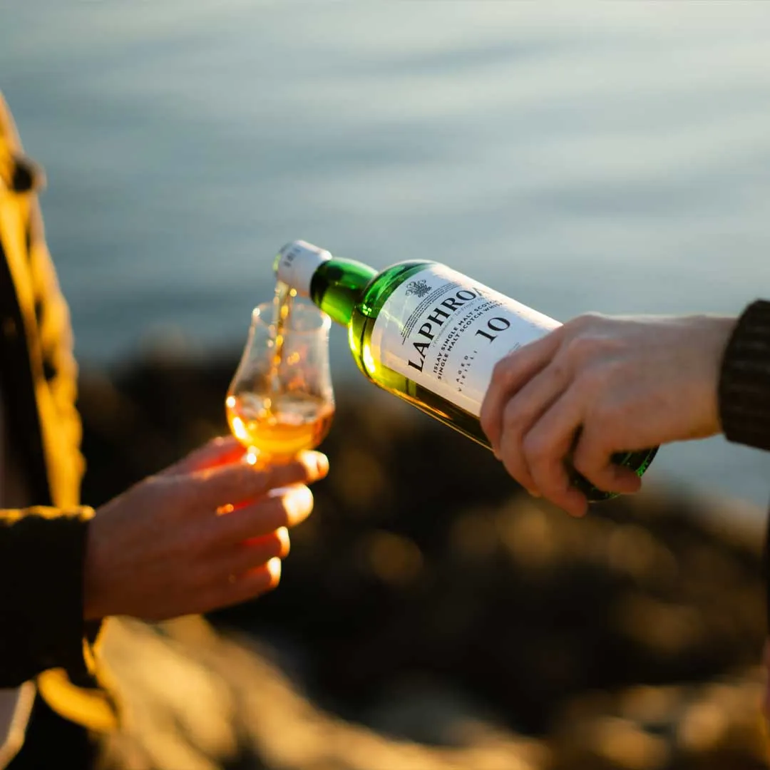 Person pouring Laphroaig 10 Year Old Islay single malt Scotch whisky into a tasting glass by the seaside.