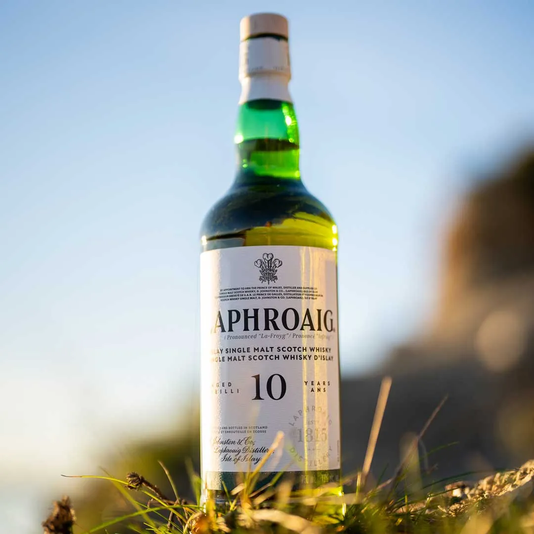 Bottle of Laphroaig 10 Year Old Islay single malt Scotch whisky standing in coastal grass with a blurred shoreline in the background.
