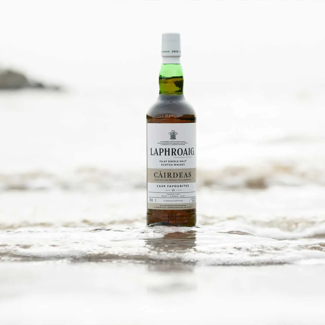Laphroaig Càirdeas Cask Favourites whisky bottle standing in shallow water on a sandy beach with a soft coastal background.