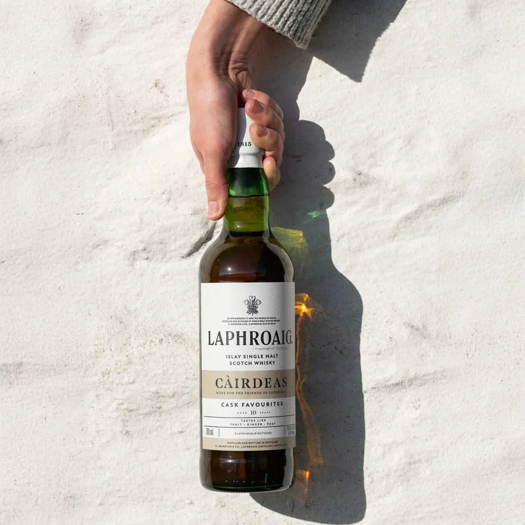Hand holding a Laphroaig Càirdeas whisky bottle against a bright sandy background with sunlight casting a warm reflection through the glass.