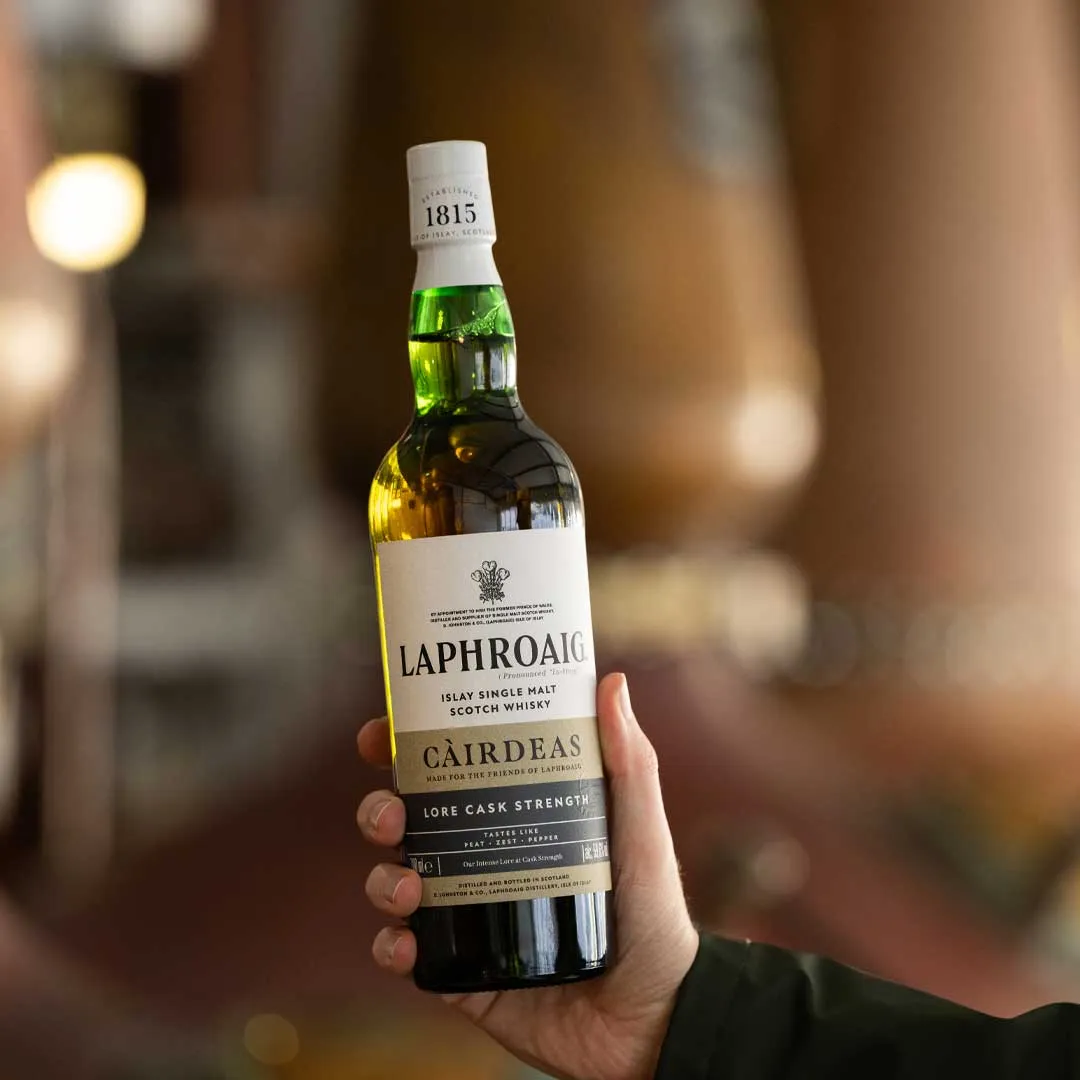 Person holding a Laphroaig Càirdeas Lore Cask Strength whisky bottle with a warm, softly blurred bar background.
