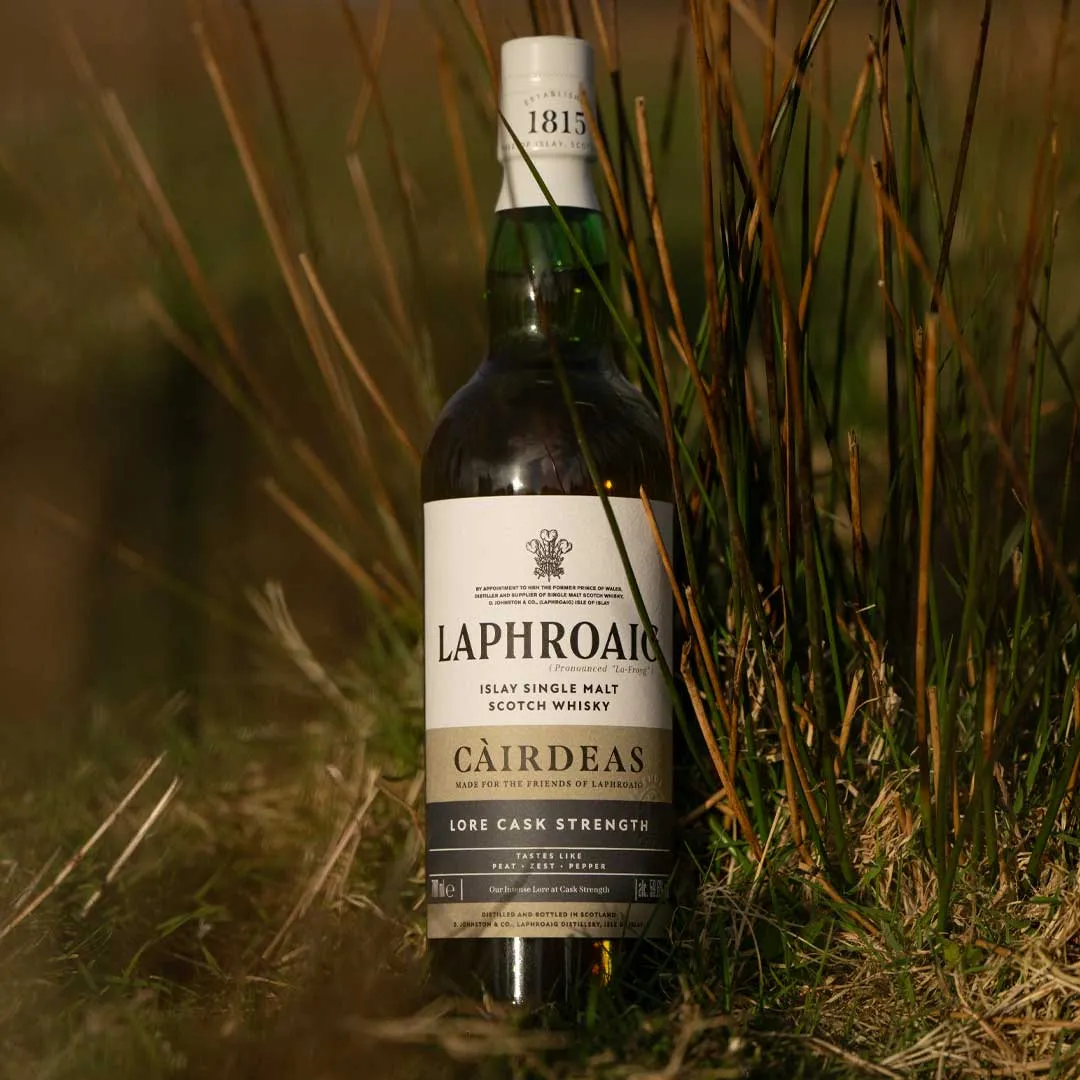 Laphroaig Càirdeas Lore Cask Strength whisky bottle placed among tall coastal grasses outdoors.