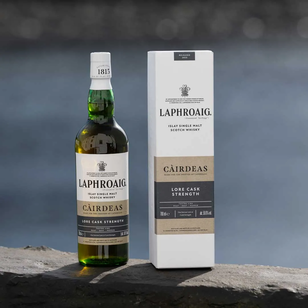 Laphroaig Càirdeas Lore Cask Strength whisky bottle next to its box packaging on a stone ledge with a blurred water background.