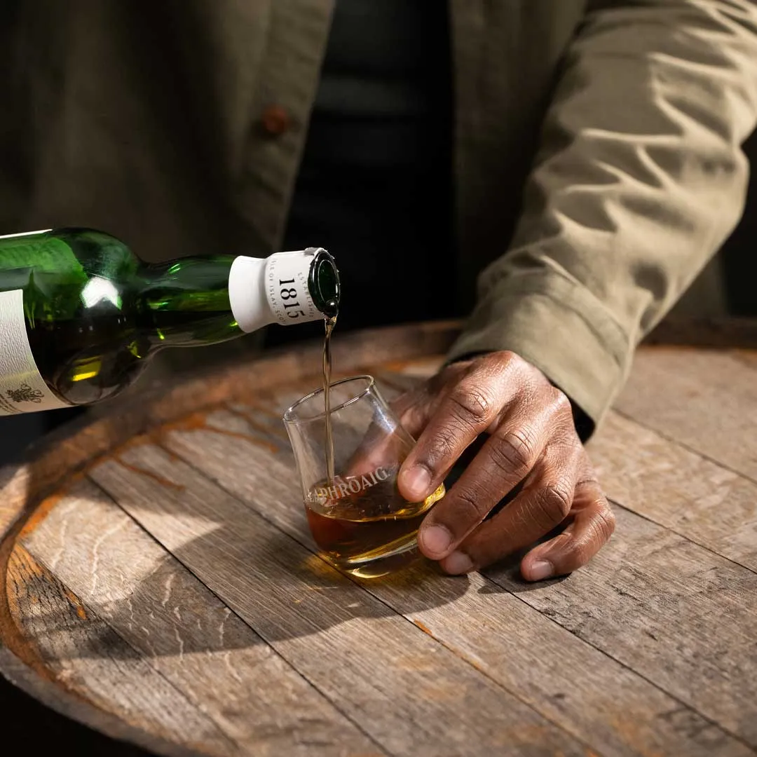 Hand pouring Laphroaig whisky from a bottle into a Laphroaig-branded tasting glass on a wooden table.