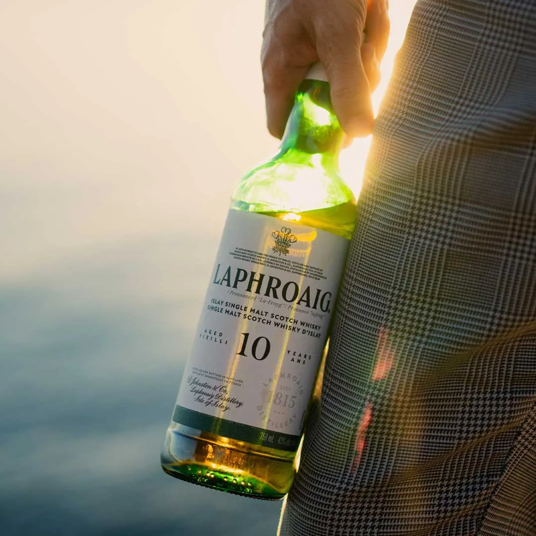 Person holding a bottle of Laphroaig 10 Year Old Islay Single Malt Scotch Whisky by the sea at sunset.
