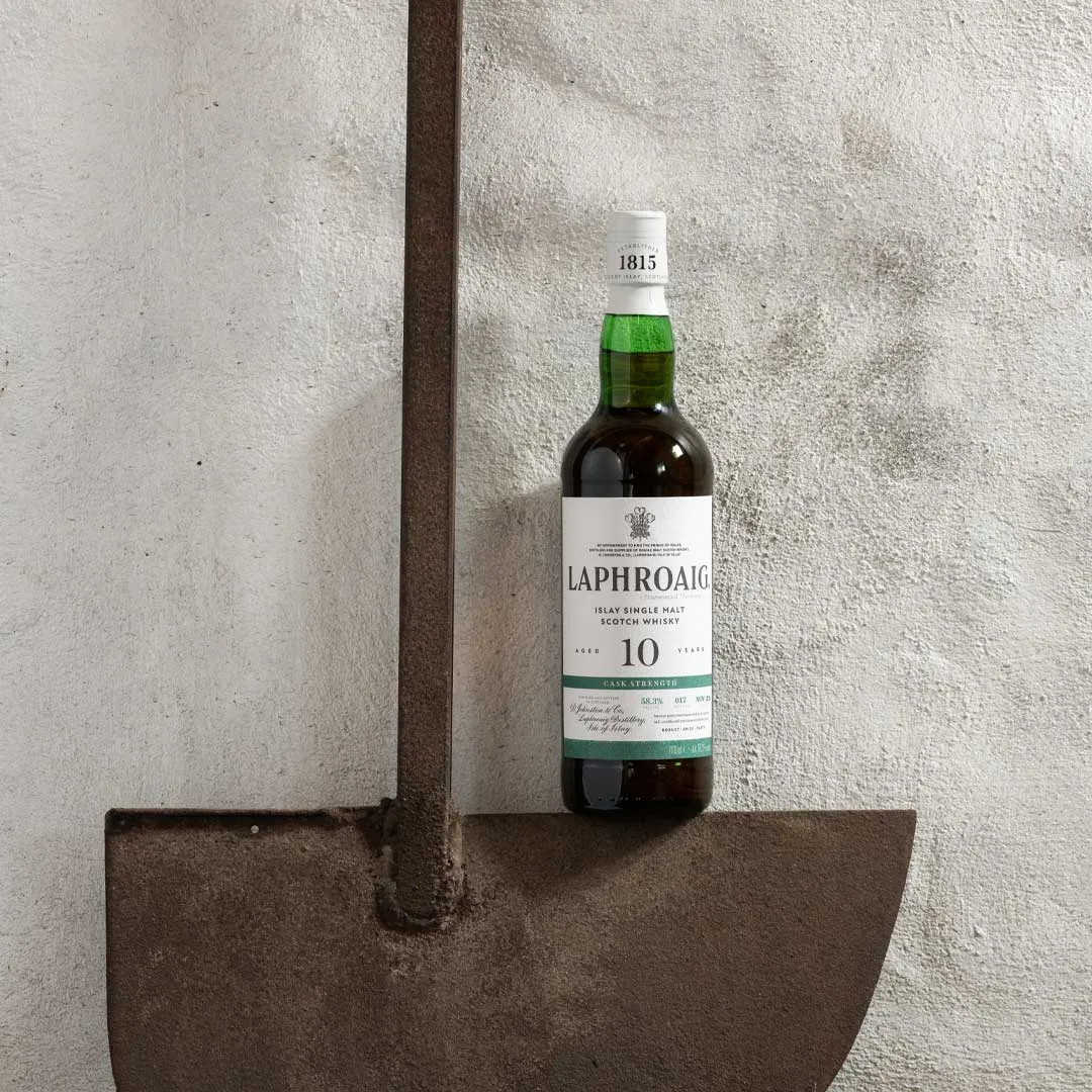 Bottle of Laphroaig 10 Year Old Cask Strength Islay single malt Scotch whisky placed on a metal shovel against a white wall.