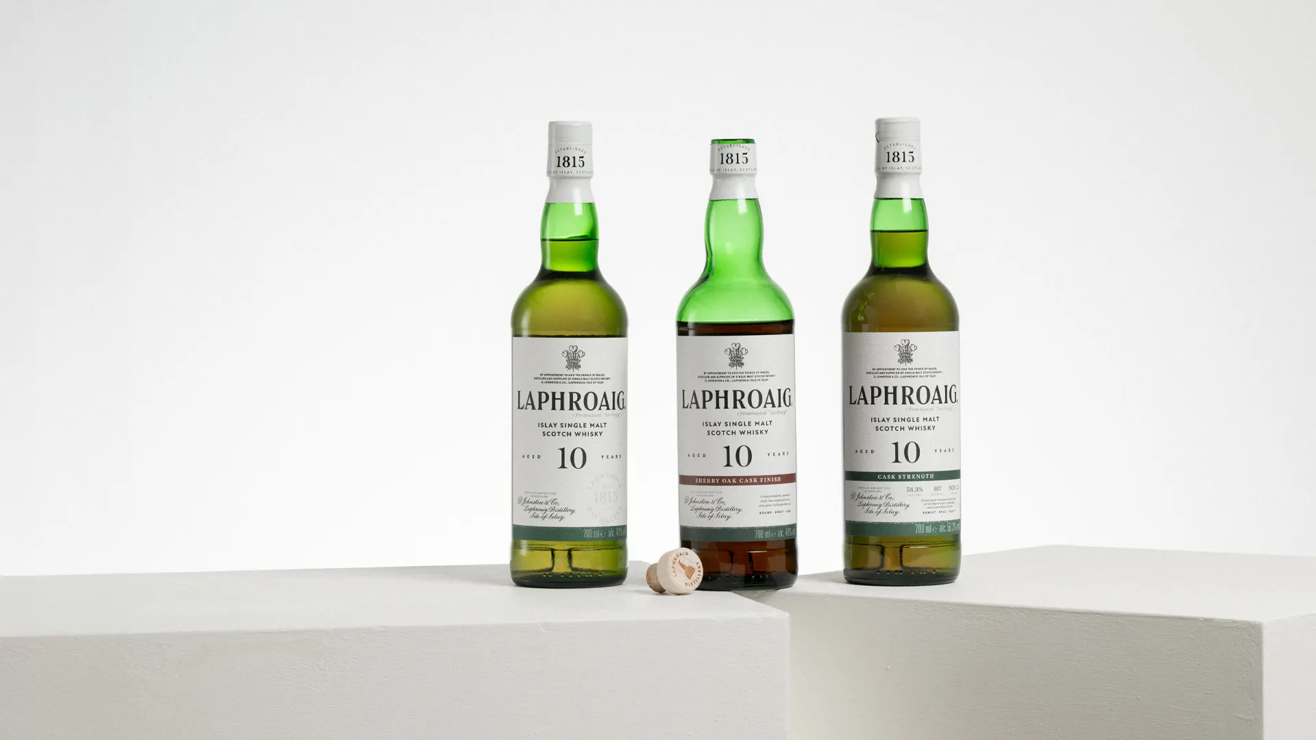 Three bottles of Laphroaig 10 Year Old Scotch whisky, including Original, Sherry Oak Finish, and Cask Strength, displayed on white plinths against a minimal background.