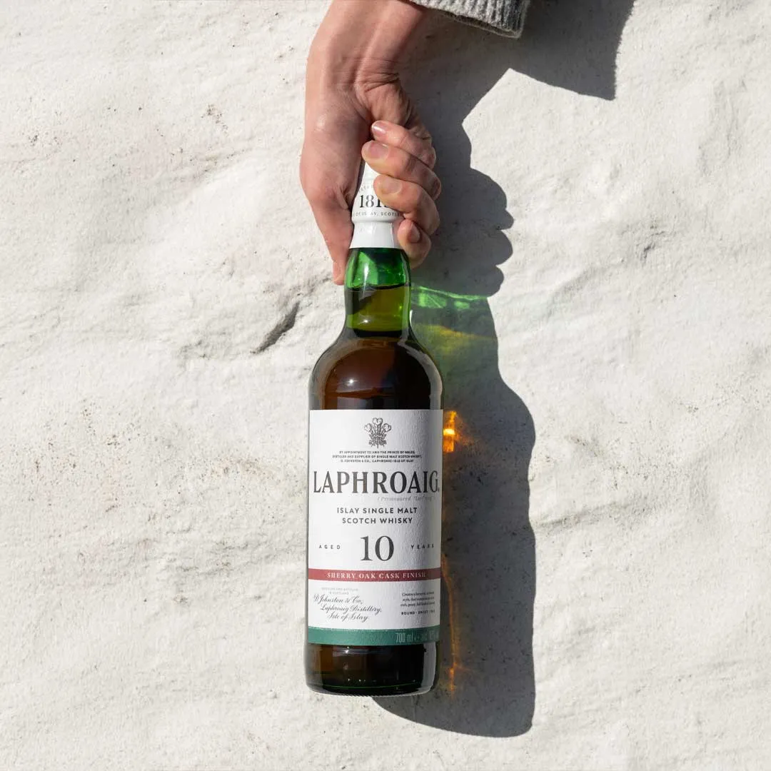 Hand holding a bottle of Laphroaig 10 Year Old Sherry Oak Cask Finish Islay single malt Scotch whisky against a white wall.