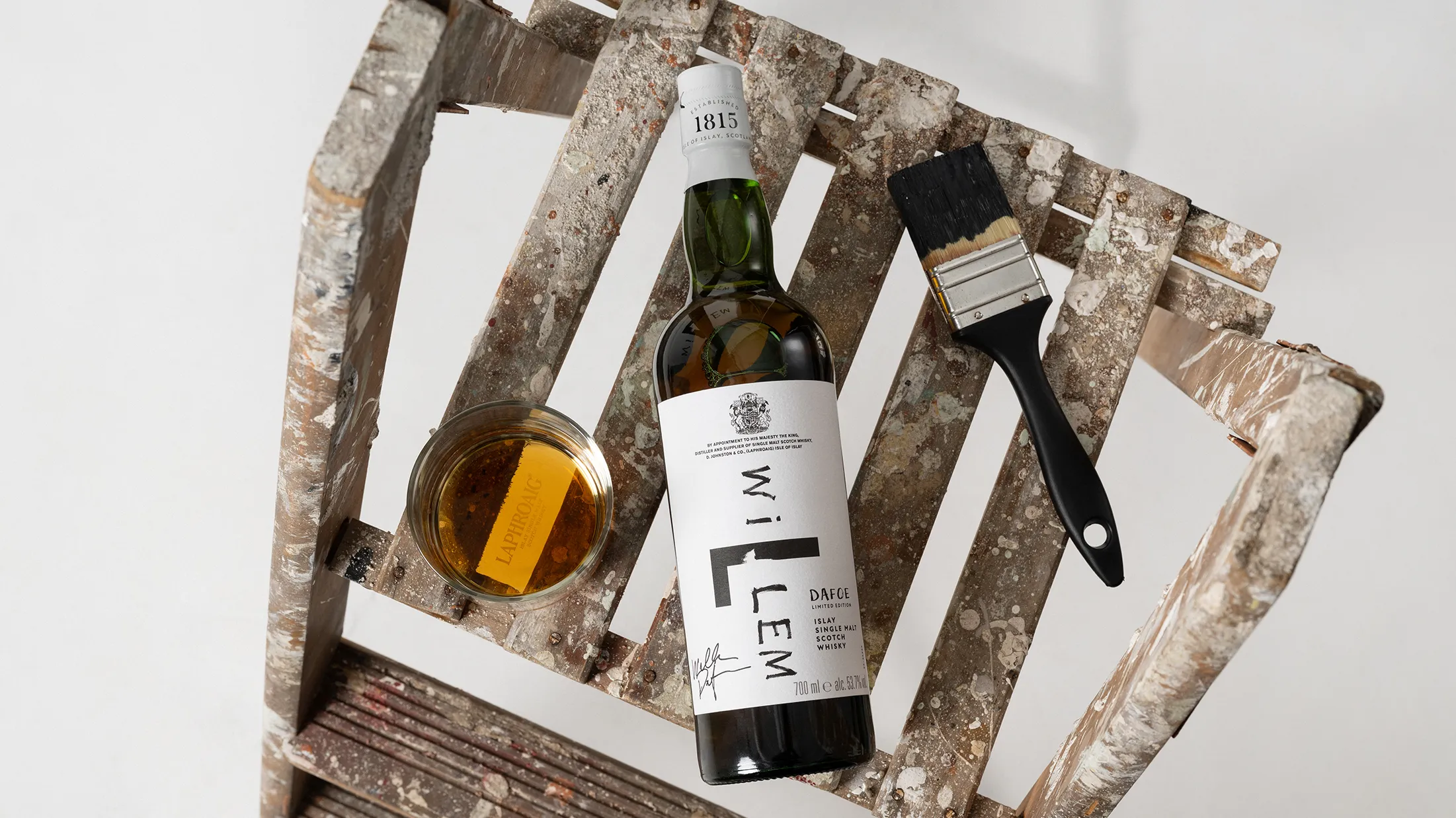 A whiskey bottle labeled "Willem," a glass of whiskey, and a paintbrush rest on a weathered wooden ladder, blending art and craftsmanship vibes.