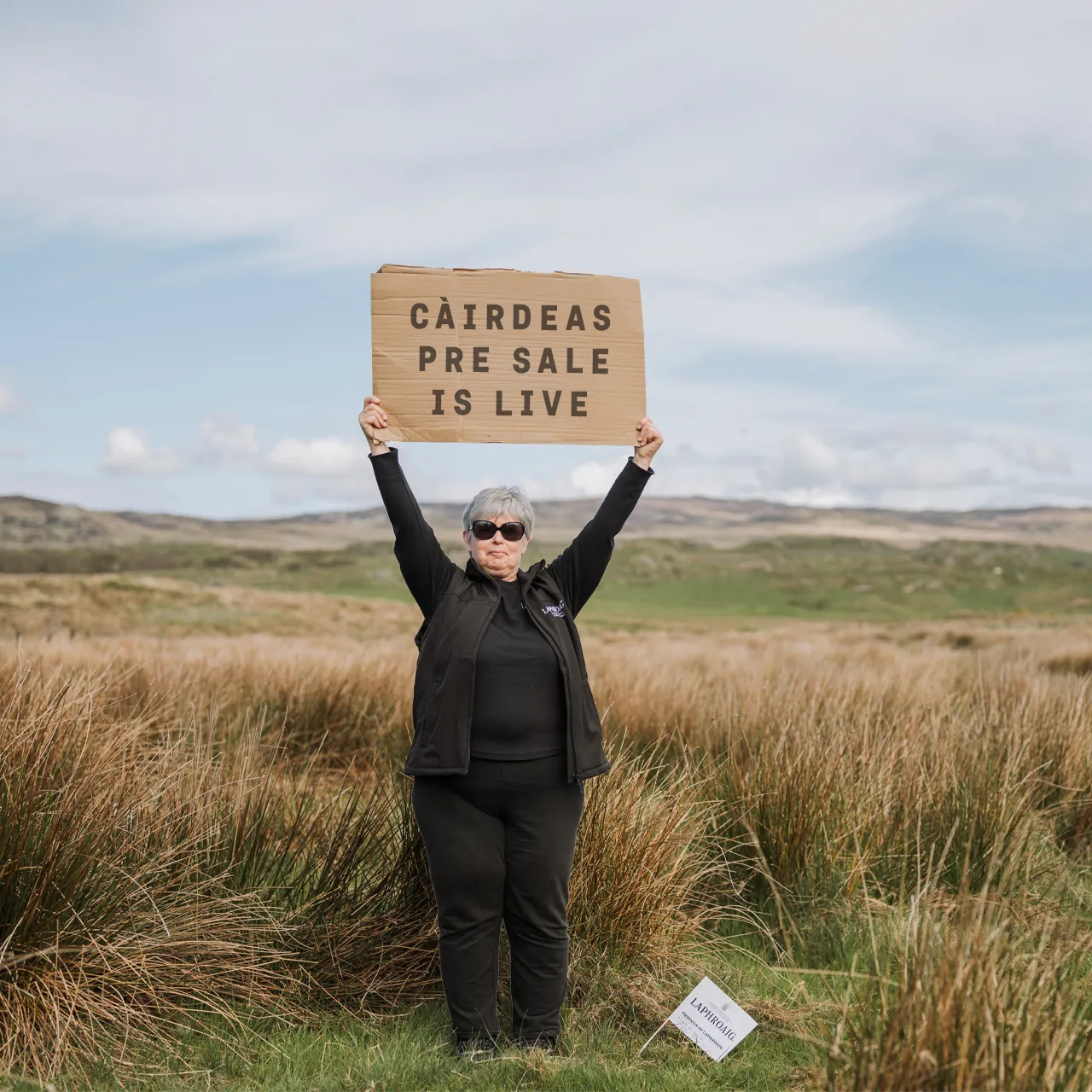 A person stands in an open grassy landscape holding a wooden sign above their head. The sign reads:  “CÀIRDEAS PRE SALE IS LIVE”  They are wearing dark clothing and sunglasses, and there’s a small sign in the grass near them. The background shows rolling countryside under a cloudy sky.