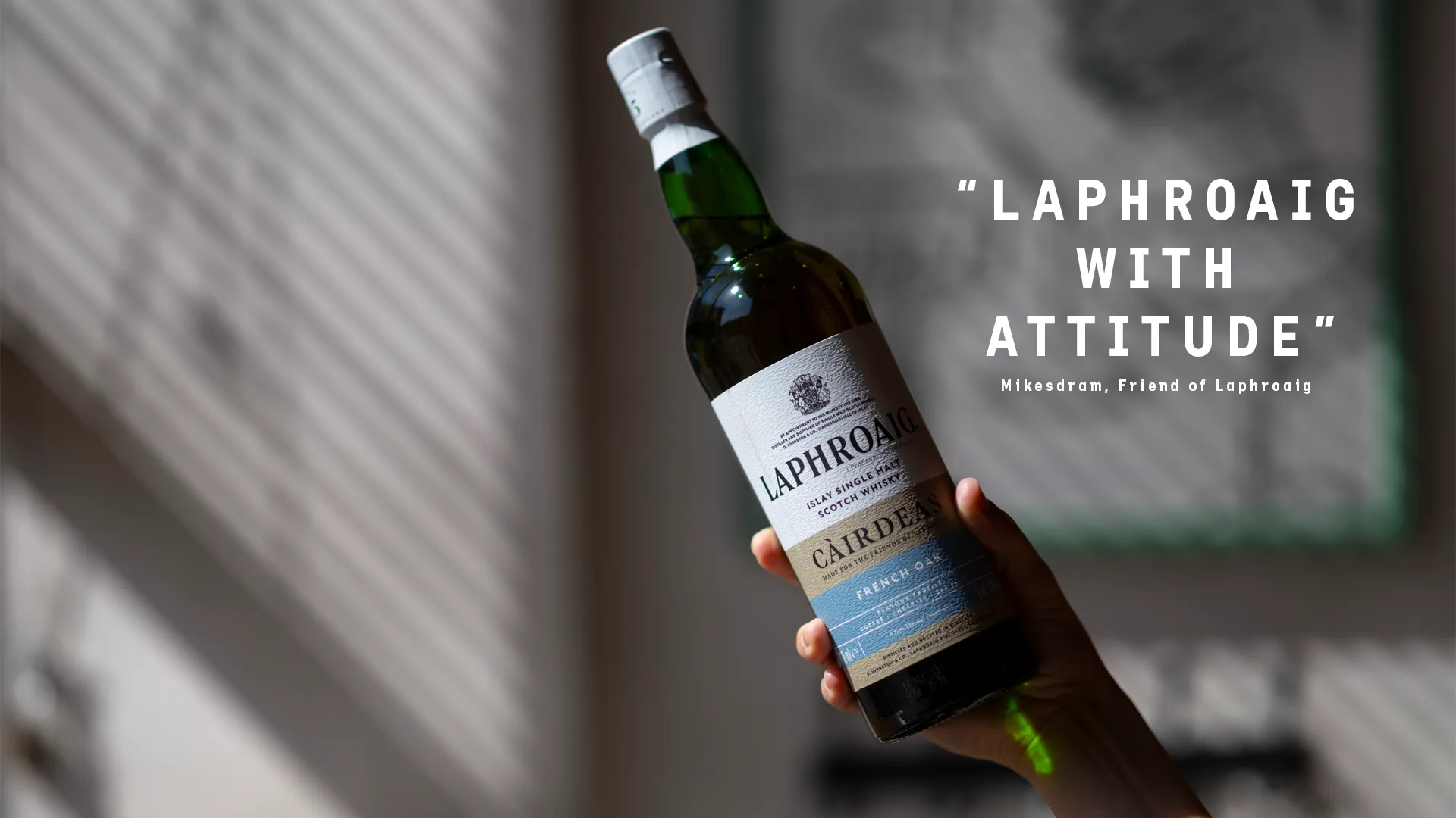 Hand holding a bottle of Laphroaig Cairdeas French Oak whisky, lit by slatted window shadows, with the words “Laphroaig with attitude” on the right.