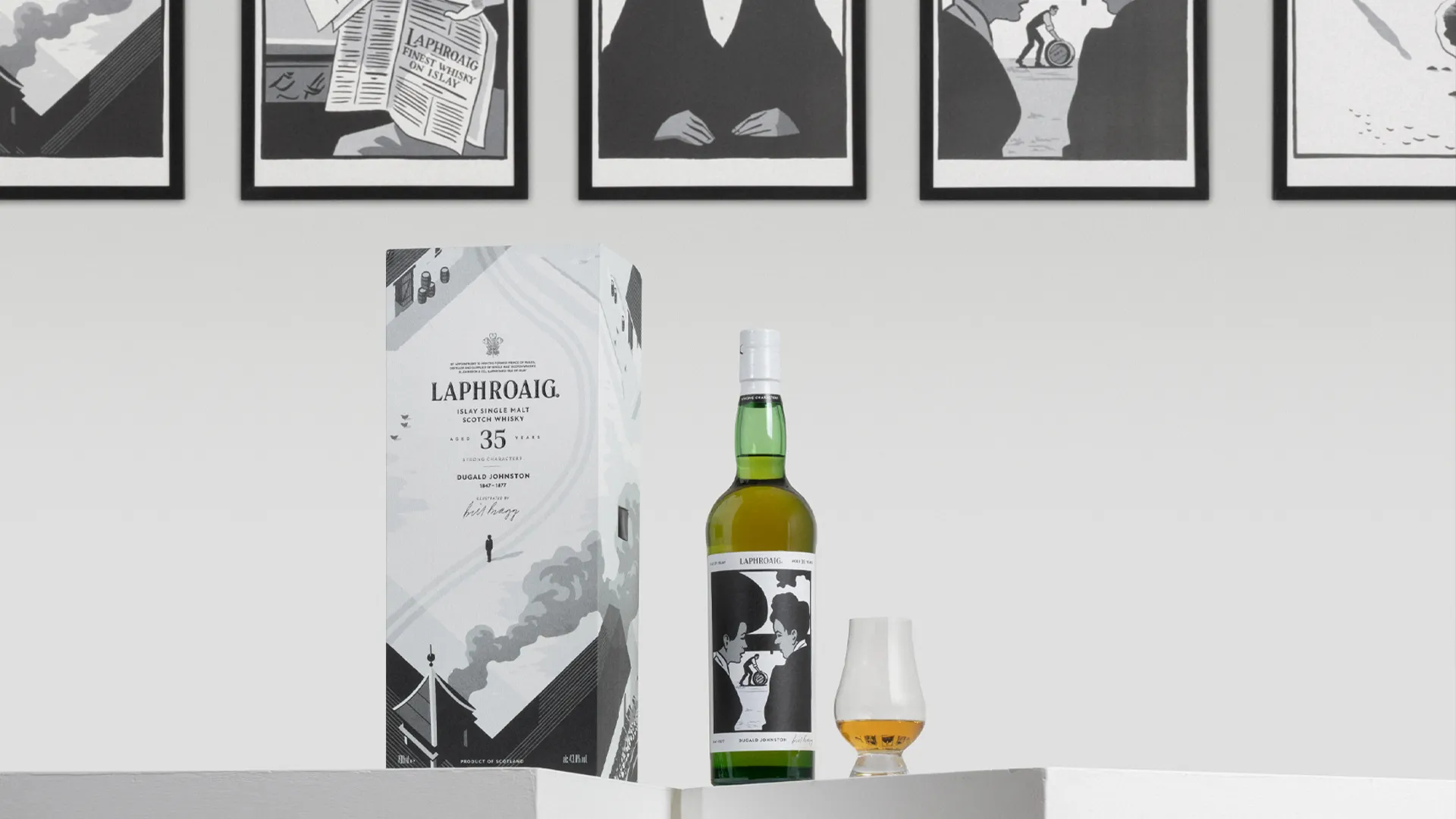 Laphroaig 35 Year Old whisky presentation featuring an illustrated box, green bottle, and a tasting glass with amber whisky on a white display surface. Framed black-and-white illustrations hang on the wall behind the products, echoing the artwork on the packaging and bottle label.