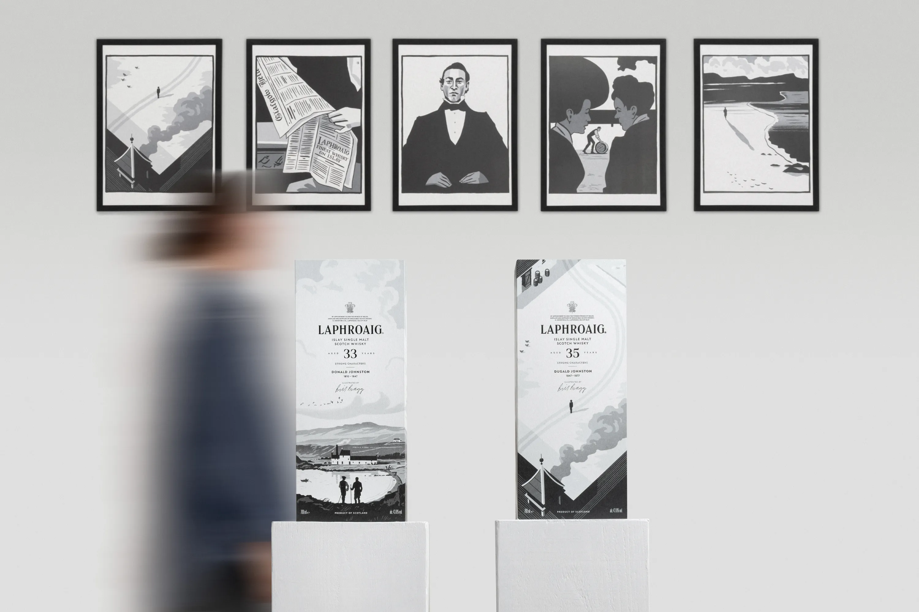 Laphroaig Strong Characters 33-Year-Old and 35-Year-Old whisky presentations displayed side by side beneath framed illustrations, with a blurred figure passing in the foreground.